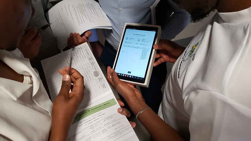 Healthcare workers in Namibia review laboratory test results on a tablet while holding patient forms and records.