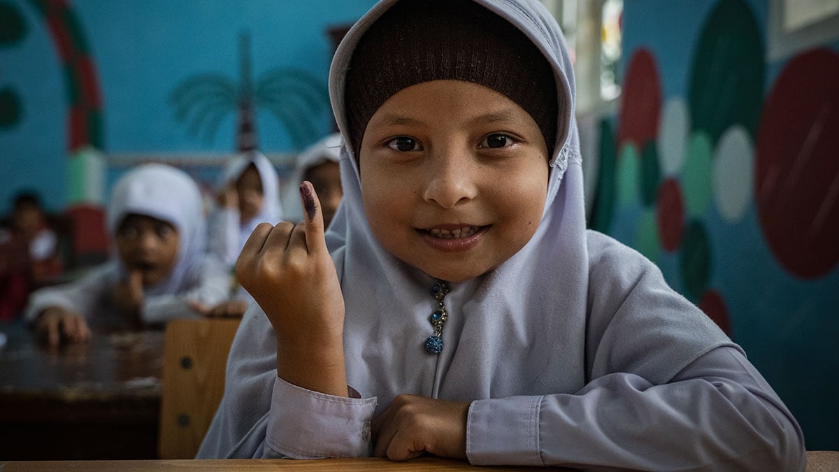 A girl in a classroom shows a mark on her finger indicating she has received an oral polio vaccine.