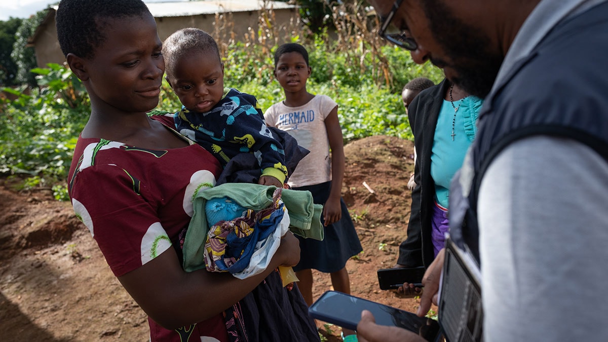 A woman holding a baby talks to a health worker tracking vaccinations with a phone app.