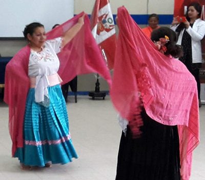 Three women dance with red scarves in a room. One woman in a white shirt looks on in the back.