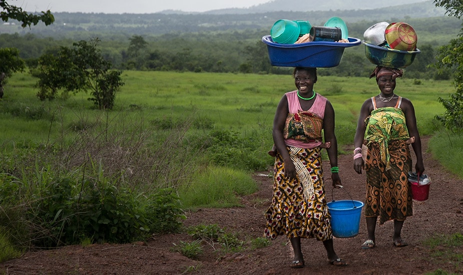Two women from Guinea carrying children on their backs and balancing laundry dishes on their heads. Photo: Scott McPherson, RTI International