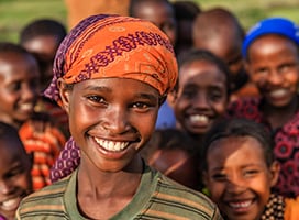 A child in a brightly colored headwrap smiles at the camera, with a group of children smiling in the background.