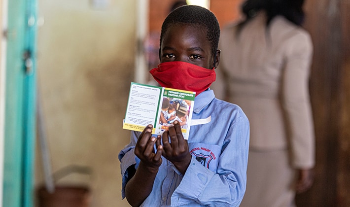 A young boy wearing a mask holds up a typhoid fever vaccination card in Zimbabwe 2021.