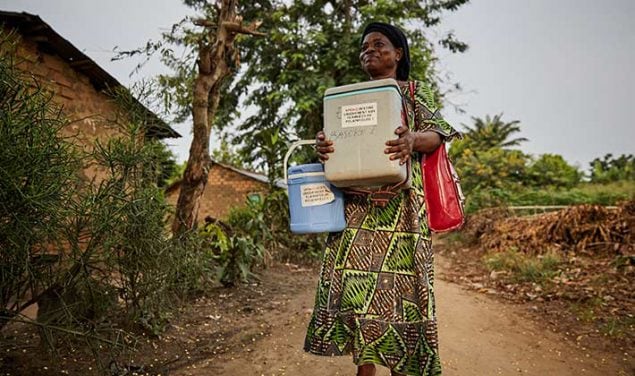 Entrega especial: uma enfermeira de vestido tradicional carrega 2 grandes refrigeradores de vacina contra poliomielite por uma estrada de terra na RDC.