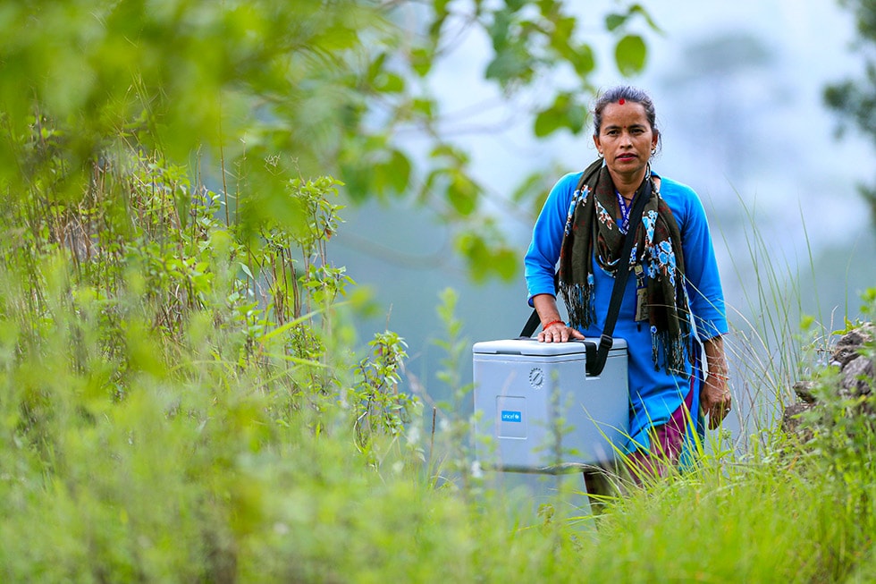 A female health worker carries a large cooler of vaccines alone through the backcountry of Nepal.