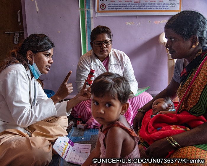 A mother with an infant and small child watches as 2 female healthcare workers conduct a routine immunization session.