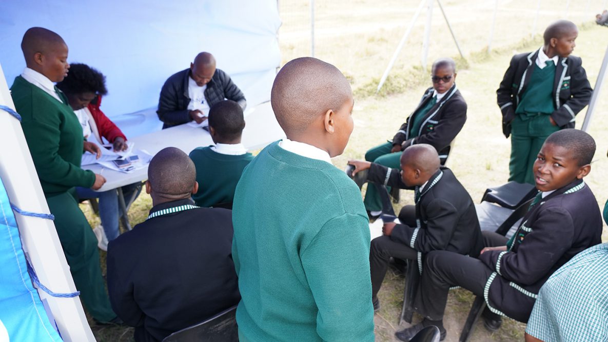 Photo of a group of students sitting inside a tent outside their school waiting to receive COVID-19 vaccines.