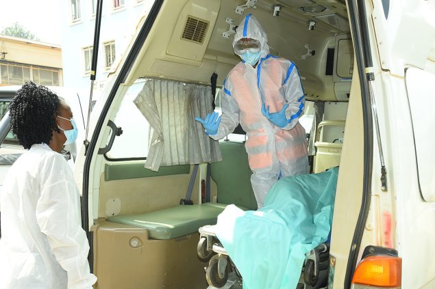 A nurse wearing a surgical mask stands next to an ambulance while talking to an ambulance worker inside the vehicle