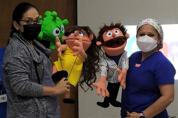 Four health workers at a clinic in Santa Ana, El Salvador stand in front of a woman who is sitting. They use puppets to explain how people expecting a baby benefit from getting the COVID-19 vaccine.