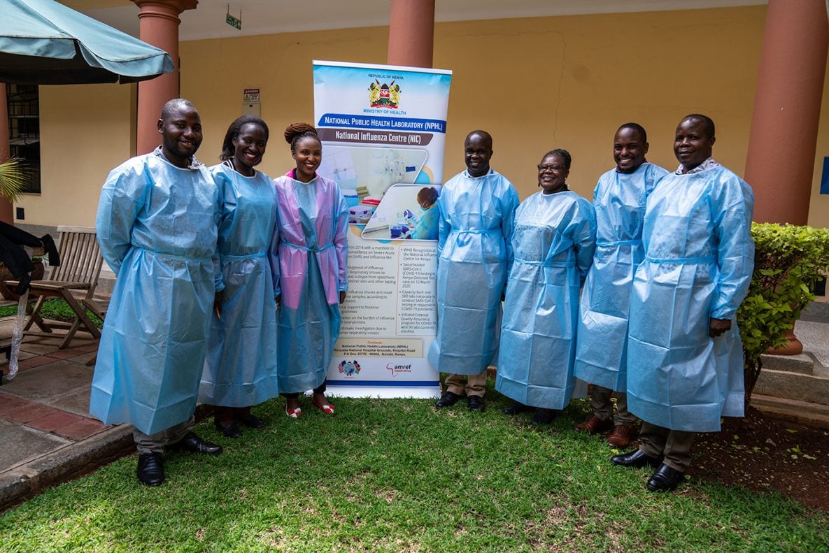 Photo of the CDC Kenya laboratory team smiling and standing outside the National Influenza Center (NIC) Laboratory. A standing sign contains the NIC logo.