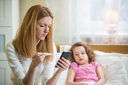 Mother checks daughter's temperature while consulting her smartphone