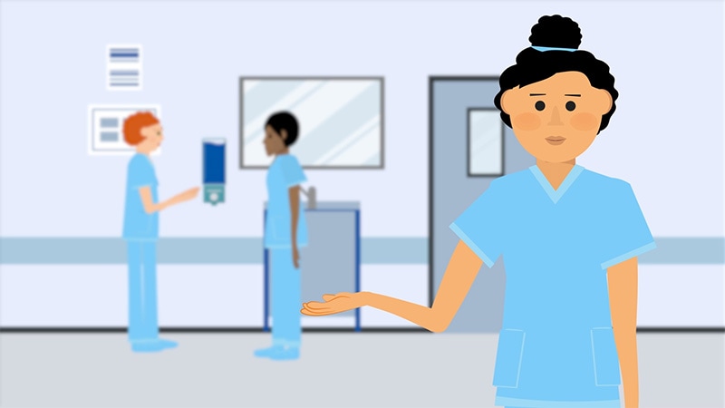 A healthcare worker in blue scrubs gestures to two healthcare workers in the background speaking to one another about proper hand hygiene.
