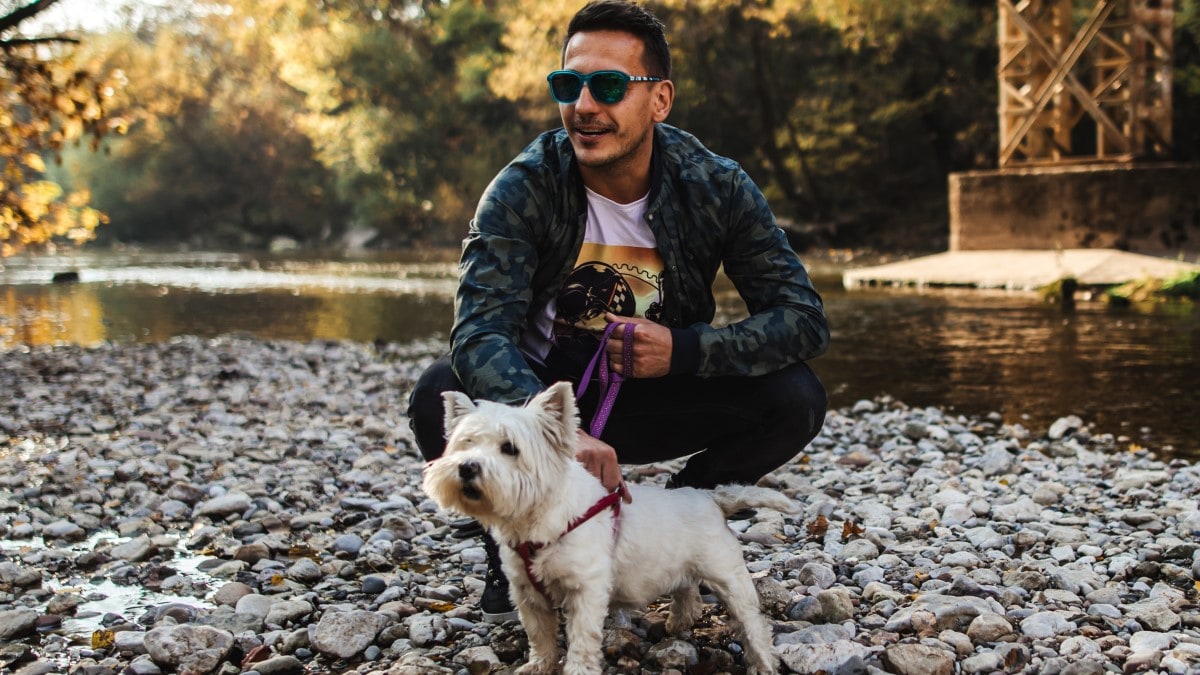 Person wearing sunglasses holding a small dog's leash on a rocky shore with a river in the background