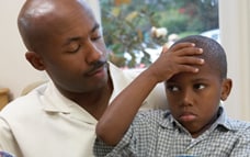 African American boy with holding his forehead while sitting on his father's lap
