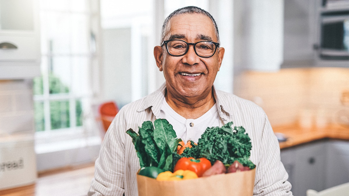 An older Hispanic man smiles while holding a bag of groceries.