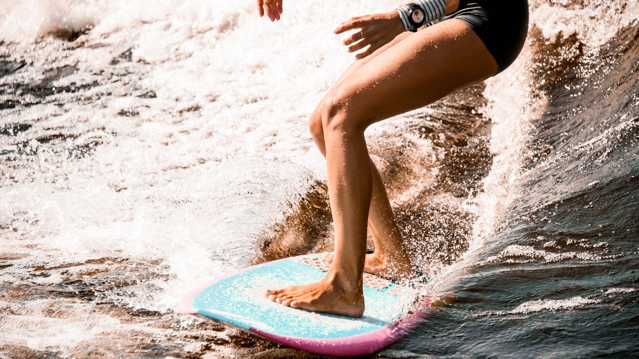 A close up picture of the legs of a person surfing with water spray behind their surfboard.