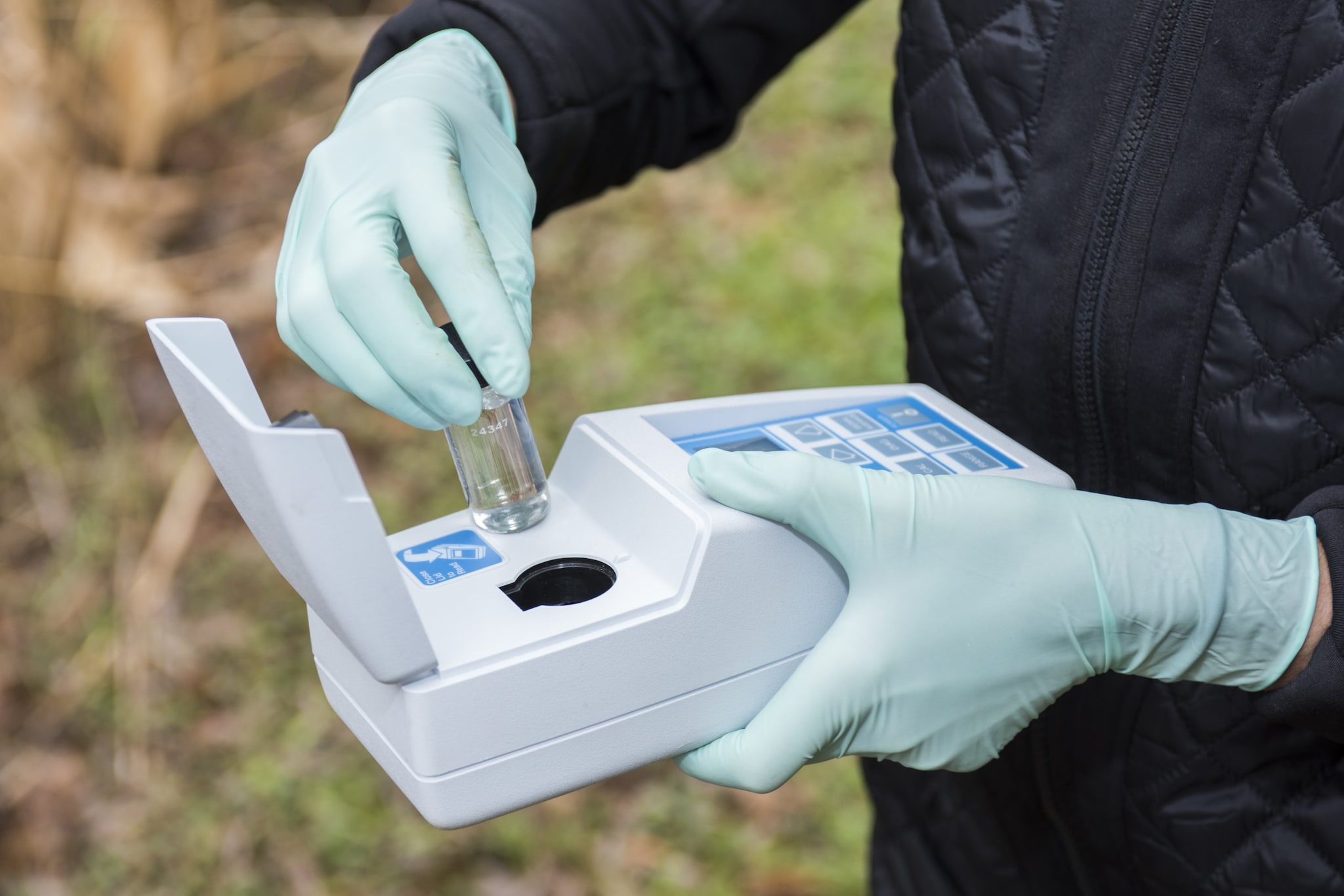 A lab scientist putting a vial of water in a handheld testing machine while outside.