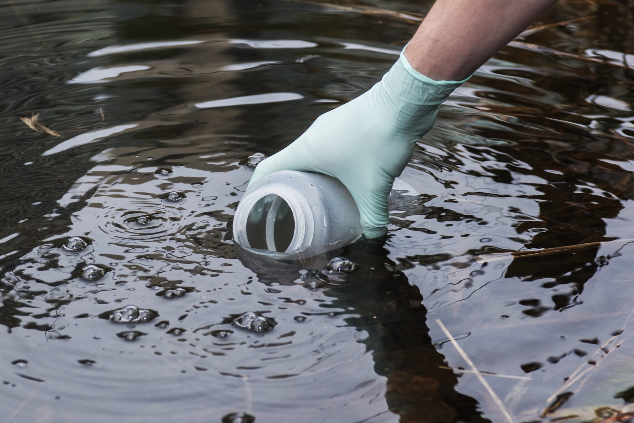 Hand with plastic glove holding a plastic bottle and submerging it in water to collect water sample.
