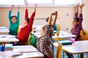 Kids sitting at their desks with their hands up in the air.