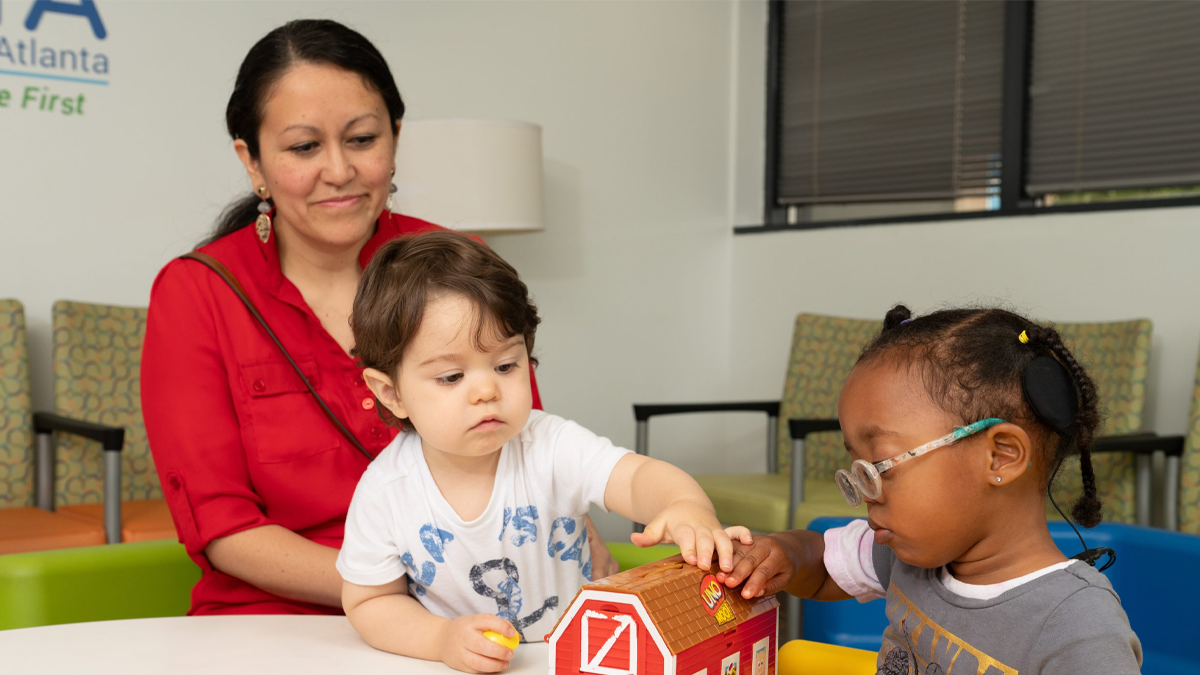 An adult supervises two children who are playing side by side while engaging in early intervention services in a clinic setting. The children are playing with a red barn toy. One child has a hearing assistive device.