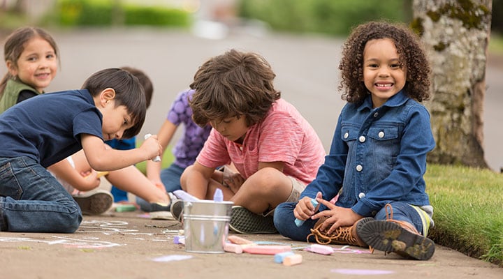 Young children sitting on the ground playing with chalk on pavement