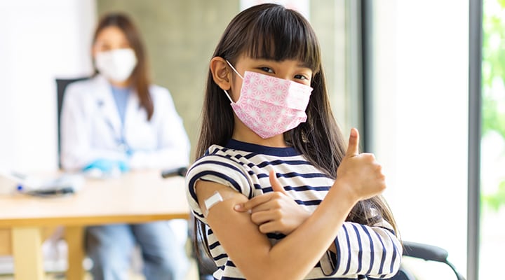 A young girl showing her band-aid on her arm and wearing a mask