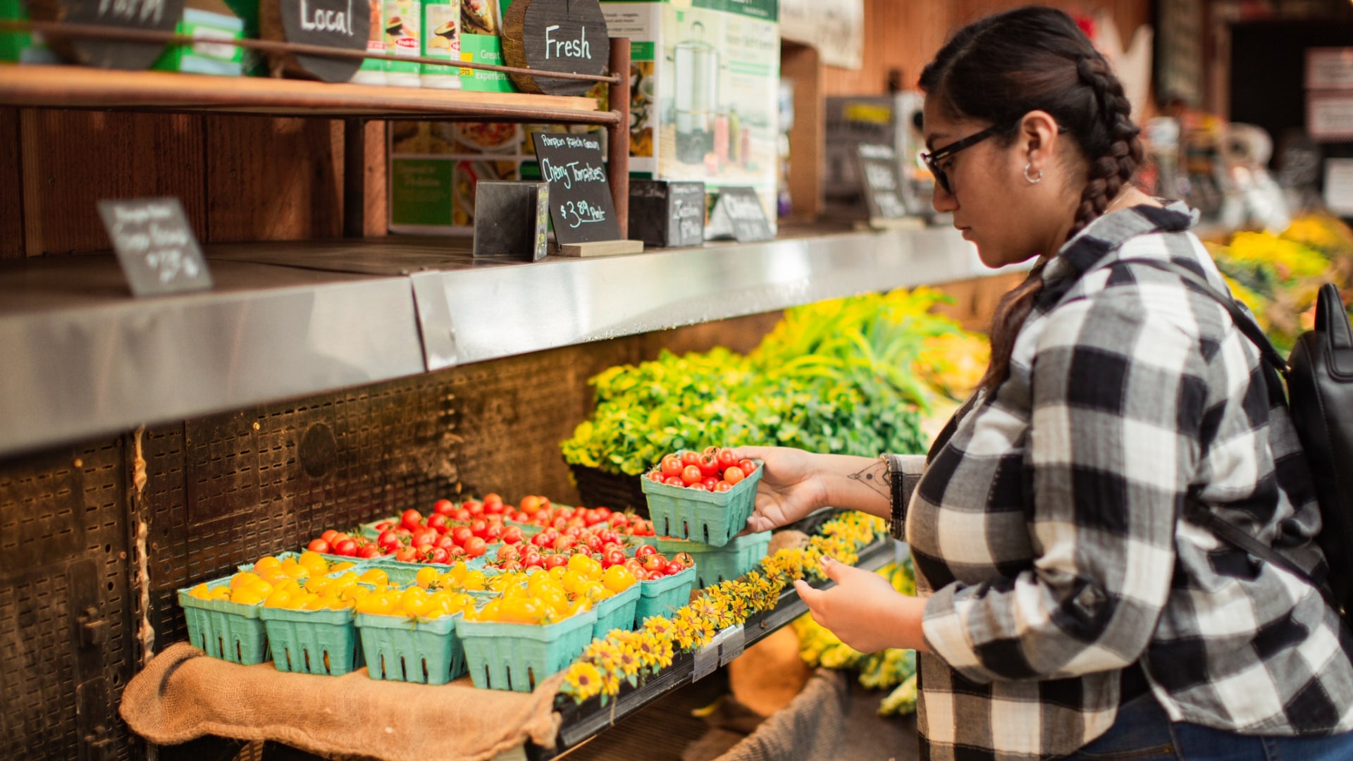 Pregnant Hispanic woman wearing a black and white checkerboard shirt, jeans, glasses with her hair braided. She is holding a carton of cherry tomatoes from the produce section at a local farm store.