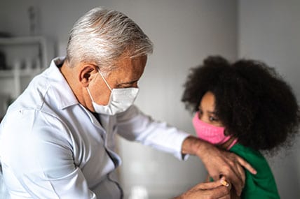 Doctor vaccinating young girl, both wearing masks