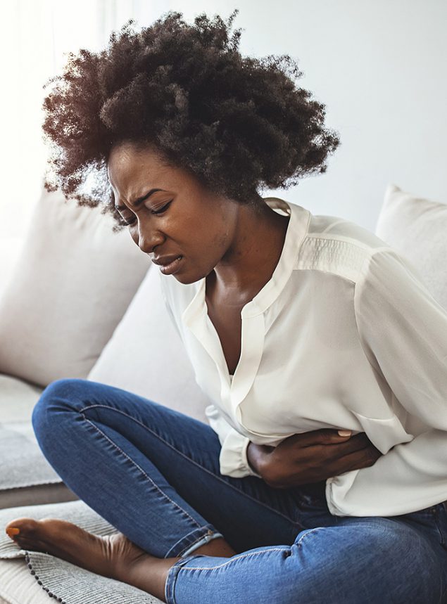 African American woman on sofa holding her stomach, experiencing abdominal pain.