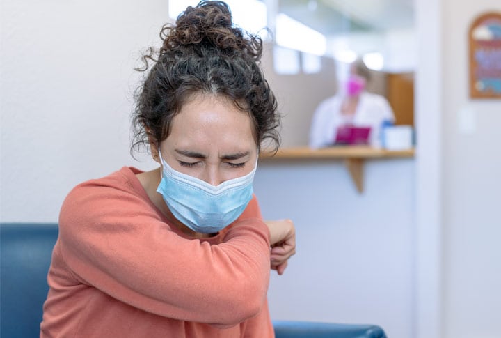 Woman wearing surgical mask sneezes into arm while sitting in patient waiting room.
