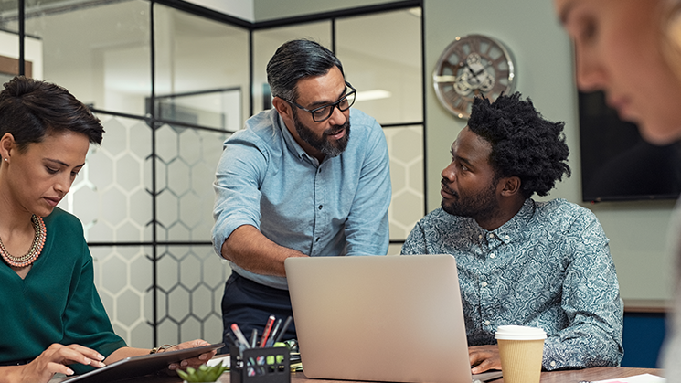 women and two men sitting around an office table working together. Two of them are discussing with an open laptop.