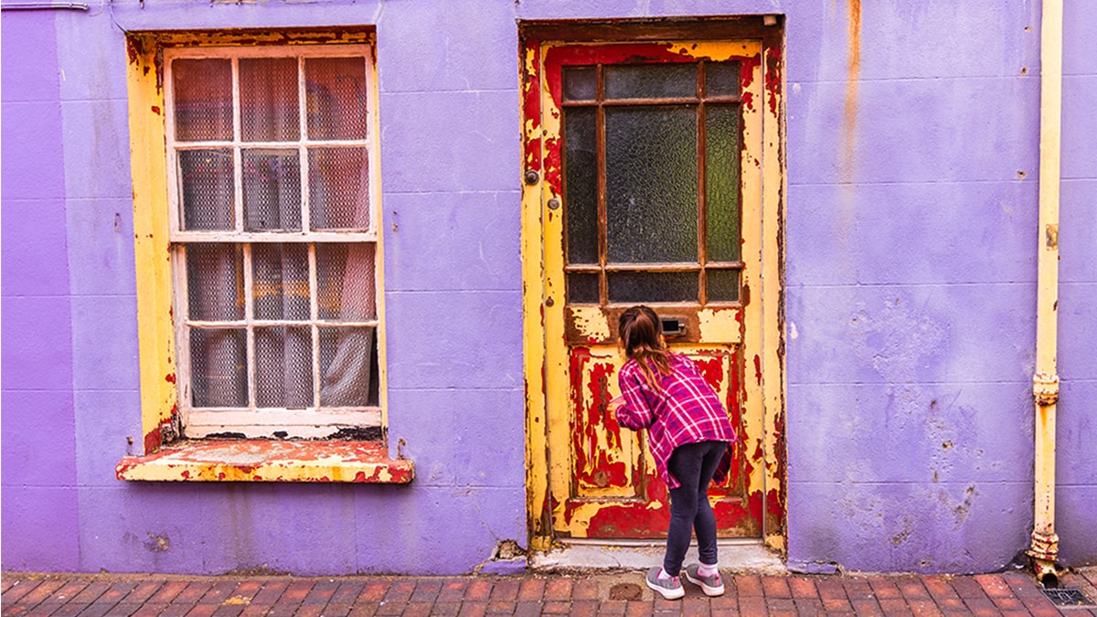 A young girl is peeping through a window in a door with peeling and chipping paint.
