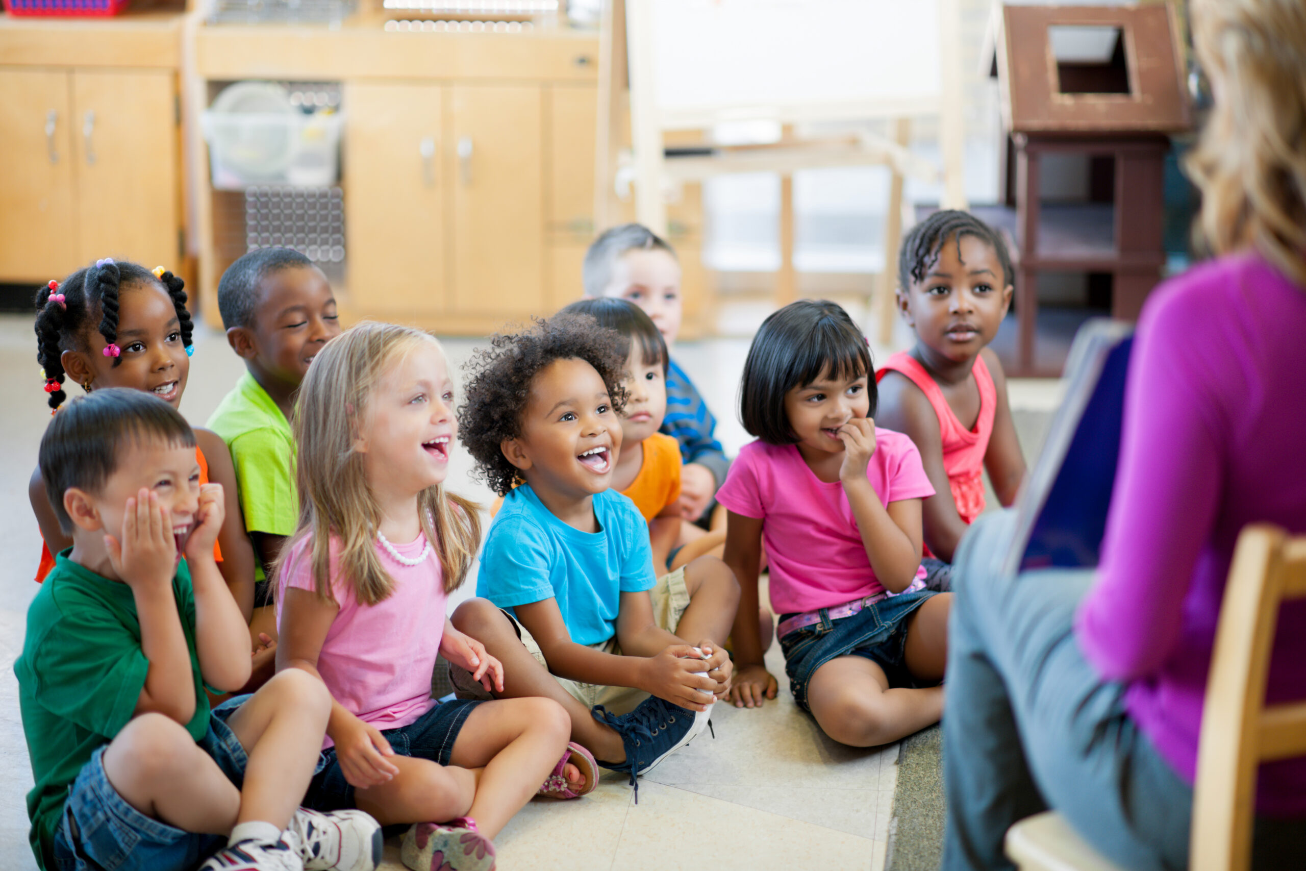 A group of nine toddlers are read to by a female teacher.