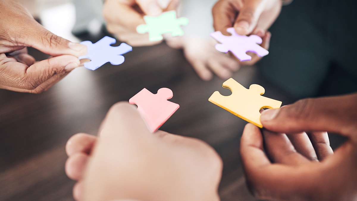 Group of diverse people around a table each holding a puzzle piece.