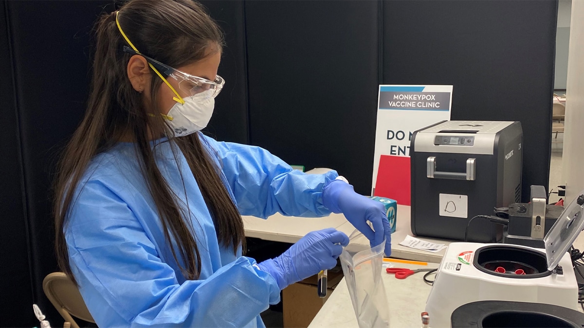 Woman in blue PPE, white mask, blue surgical gloves and lab goggles bagging a lab specimen.