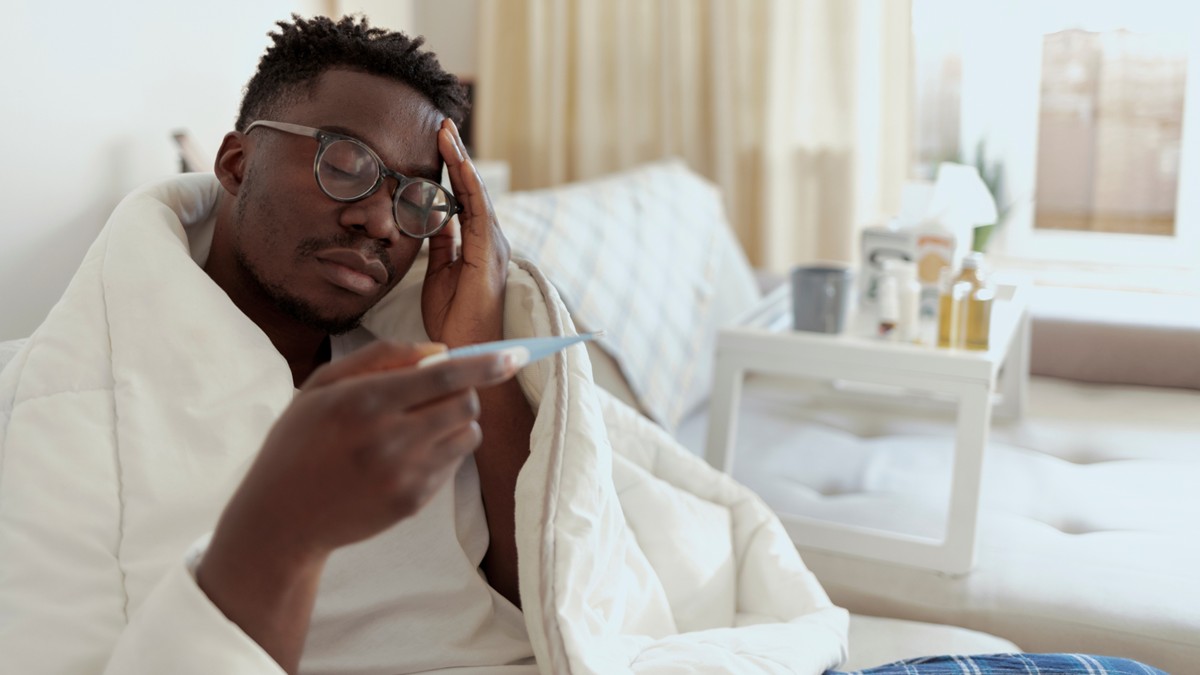 Man suffering from fever on a couch looks at thermometer.