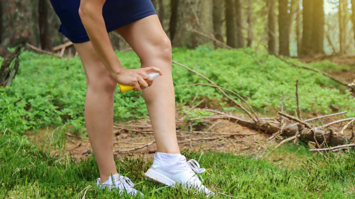 A woman sprays repellent on her left leg in a wooded area to prevent bites.