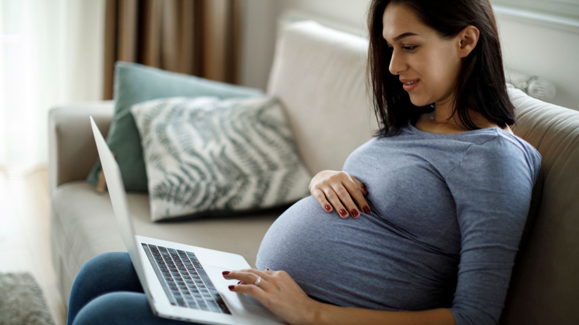 Pregnant woman sitting on a sofa and using a laptop at home.
