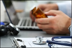 The figure above is a photograph showing a medical professional holding a bottle of pills while looking at a laptop.