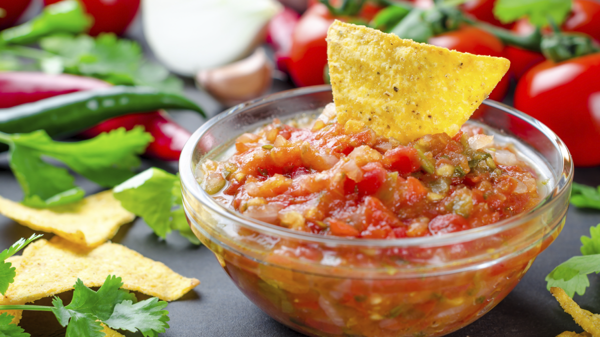 The image shows a chip in a bowl of salsa surrounded by salsa ingredients.
