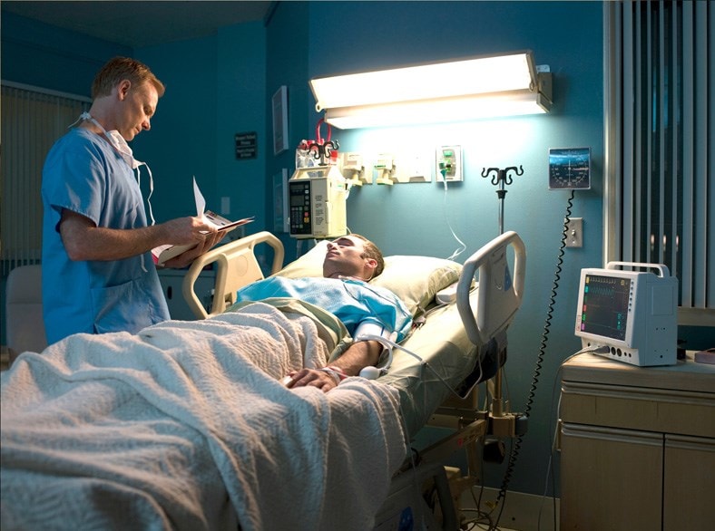 Man lying in bed at a hospital, and a doctor reviewing notes on a clipboard standing by the bedside.