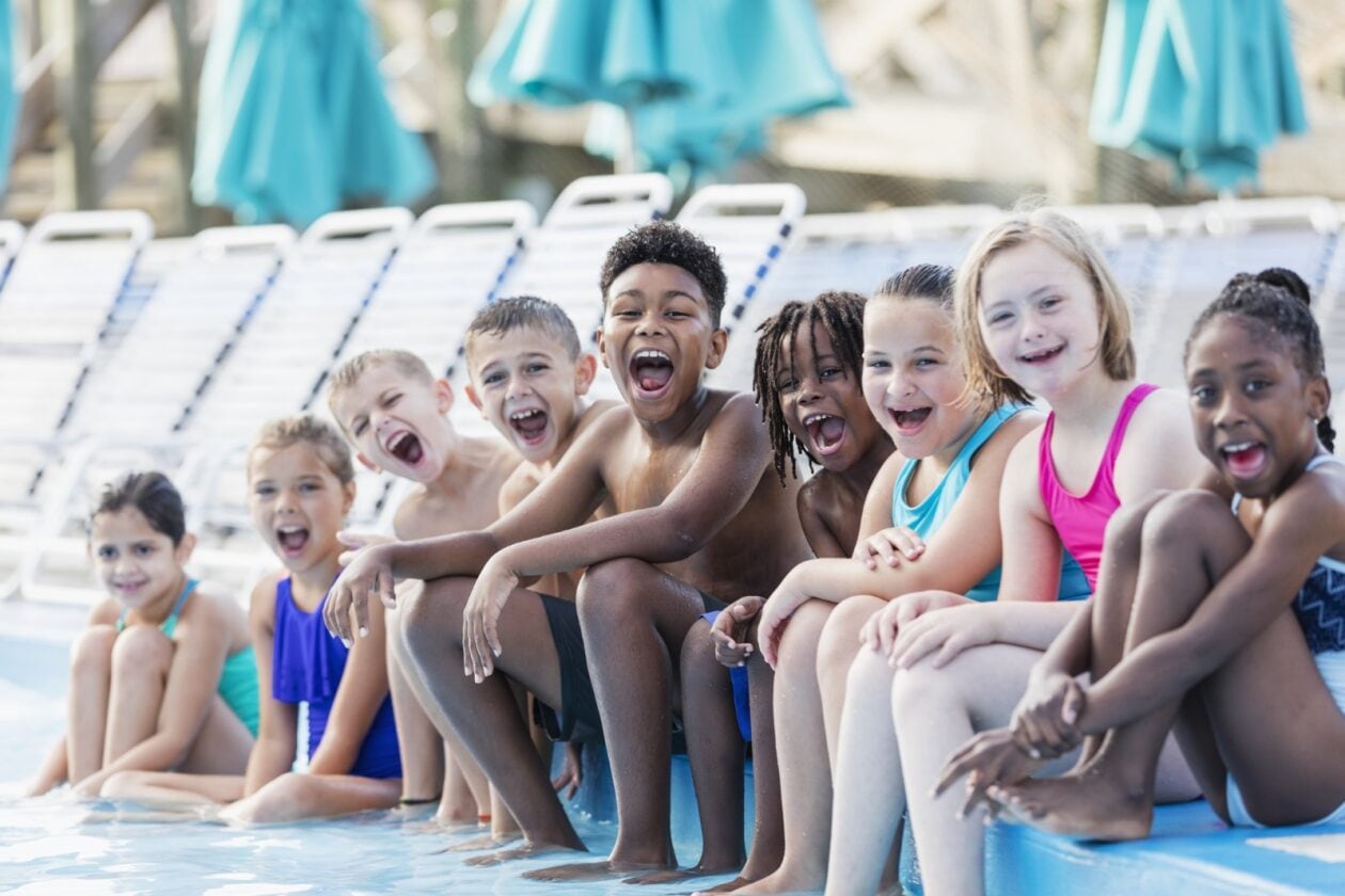 Kids of different ethnicities sitting together laughing on the side of a pool.