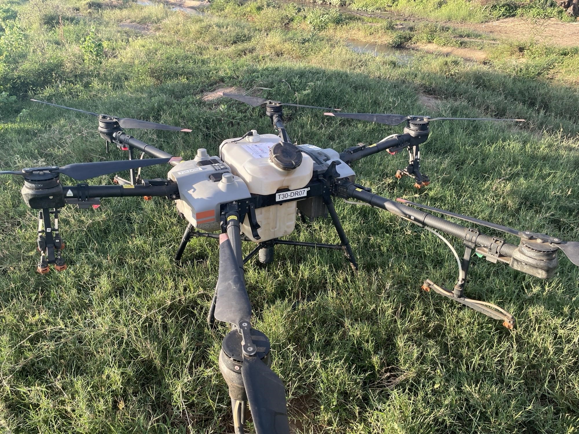 A large agricultural drone equipped with a liquid tank sits on grassy ground near a wetland or field.