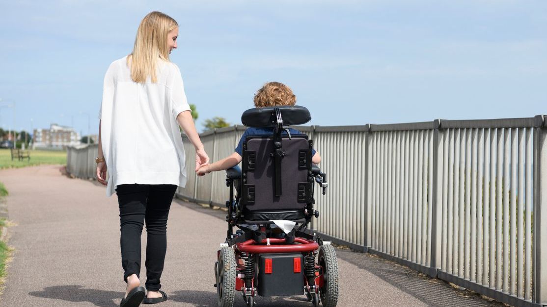A mom holds hand and walks with her son in a wheelchair.