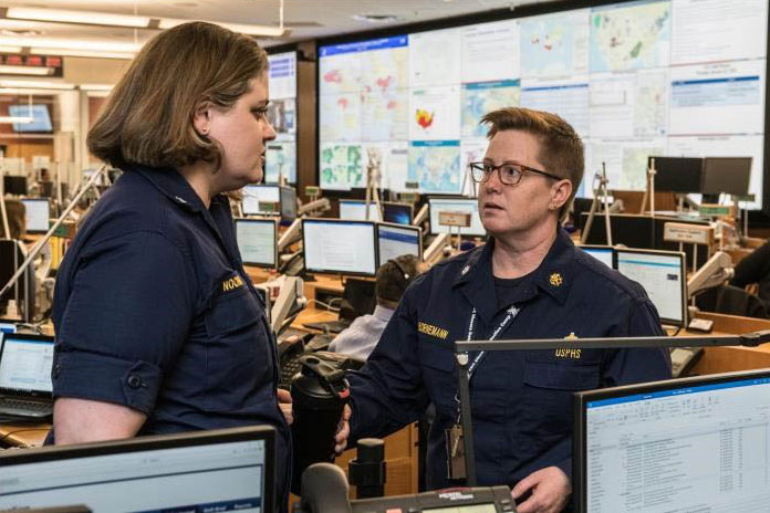 areas-study-public-health3 Two women talking in a room with many computer screens and televisions monitoring news