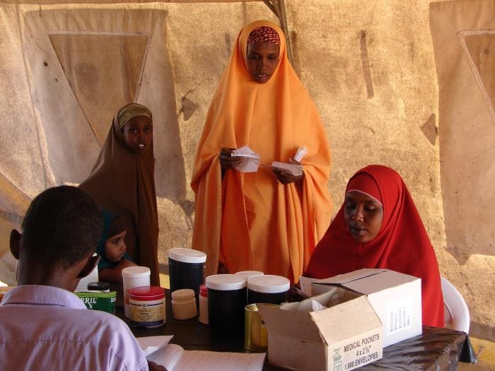 This 2007 photo, depicted the pharmacy in the Dadaab refugee camp, located near the Somali border in Kenya. Several persons are standing in a large open tent. On one side of a large table with a person working as the pharmacist providing medication.