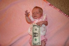 Baby Brenna dressed in doll’s clothes, half-covered by a 1- dollar bill