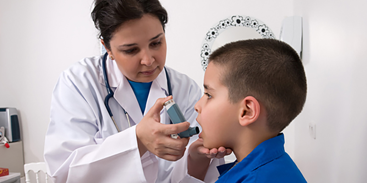 Doctor helping a boy with his asthma inhaler Image depicting a doctor preparing to use an inhaler on a young, asthmatic boy. The doctor’s hand is on the child’s chin and the child’s mouth is open to use the inhaler.