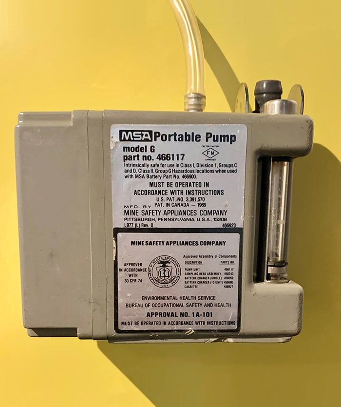 The MSA Portable Pump Model G is a grey metal box, about 5 inches by 4 inches. The pump contains a glass sample tube on the front right corner. It has a sticker that shows the logo of the USA Department of Health, Education, and Welfare.