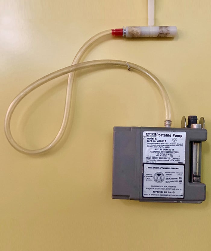 The pump has a long clear plastic tube sticking out of it that connects to a white plastic clip that would be attached to a shirt lapel. The air samples are pumped in through this tube to the sample container.
